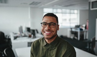 A young man wearing glasses and a button-down shirt smiling after receiving his customized dental implant in Burlington